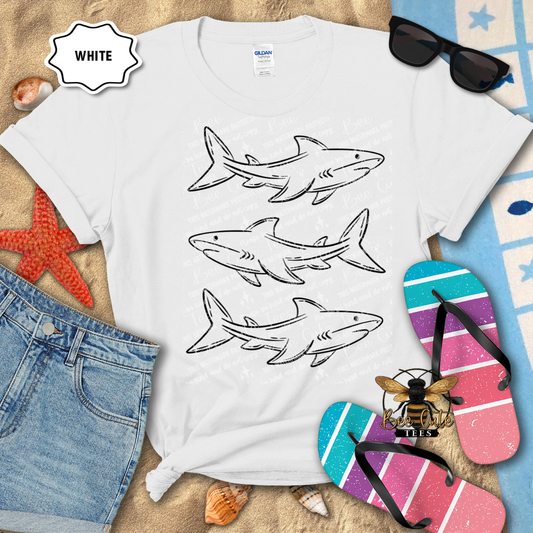 White t-shirt with shark designs on a sandy surface with beach accessories.