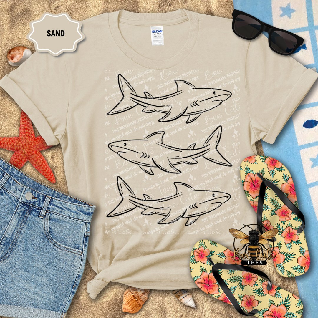 Beige t-shirt with shark designs on a sandy background with flip-flops and sunglasses.