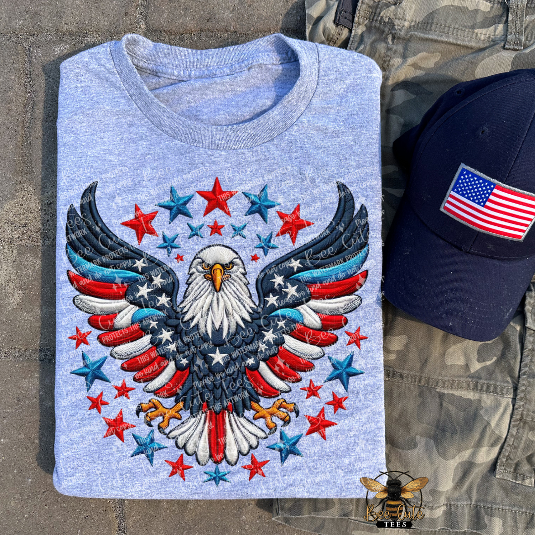 Grey t-shirt with an eagle and star design, next to a cap with an American flag patch on a stone surface.