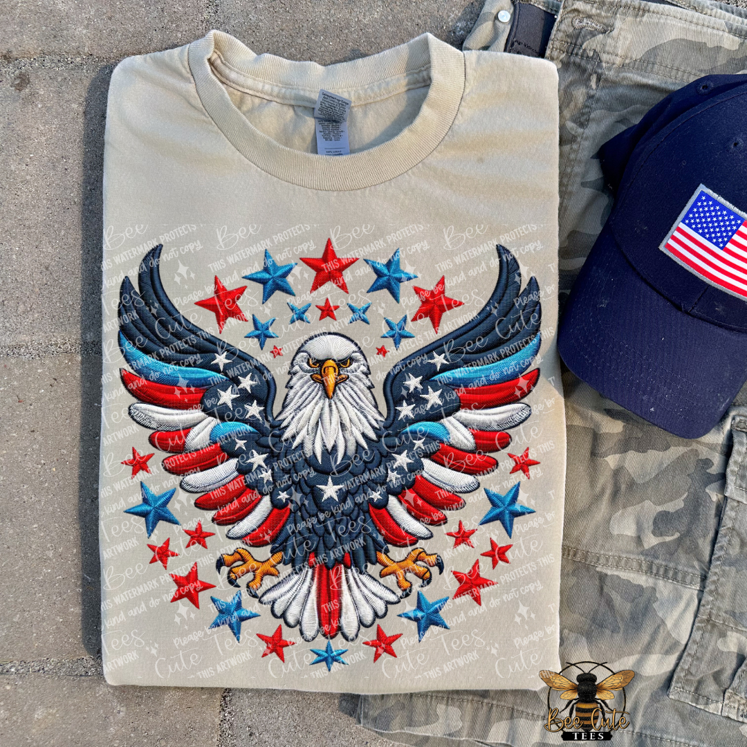 Beige t-shirt with a patriotic eagle design on a concrete surface with a cap and camouflage pants.