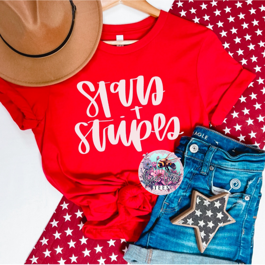 Red knotted t-shirt with a white “Stars + Stripes” script design, styled flat lay with denim shorts, a tan hat, and patriotic star-patterned accessories.

