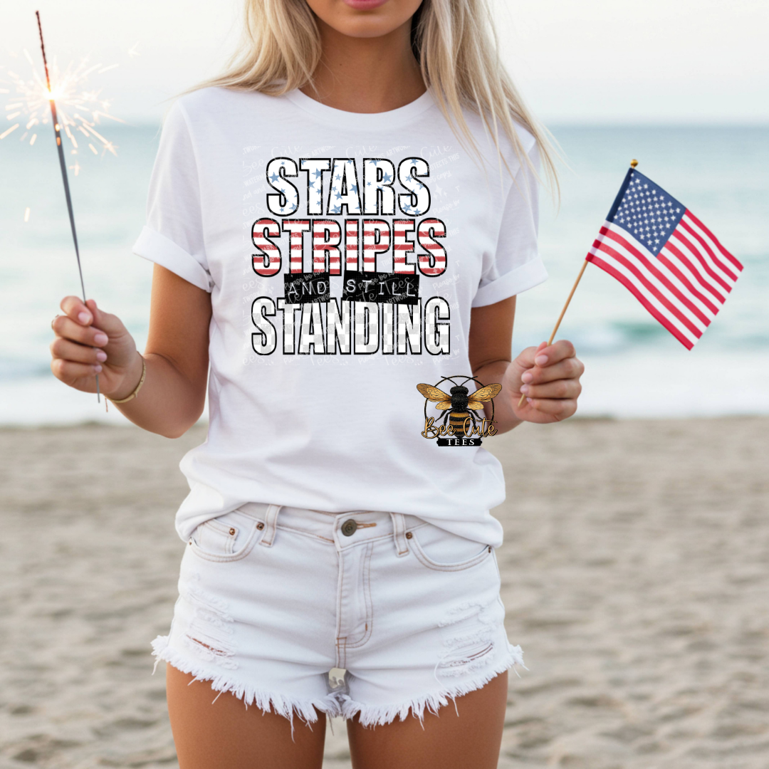 Person wearing a white t-shirt with 'Stars Stripes and Still Standing' text on a beach.