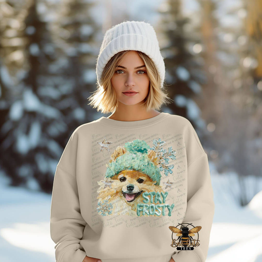 Woman wearing a beige sweater with a dog graphic and 'Stay Frosty' text in a snowy forest setting.
