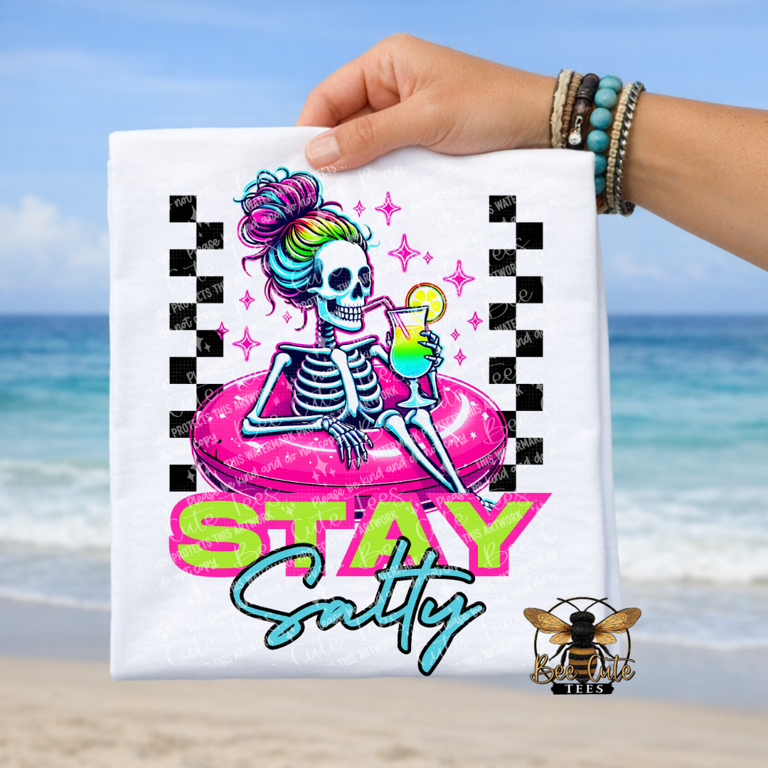 T-shirt with a skeleton design and 'Stay Salty' text held by a person on a beach.