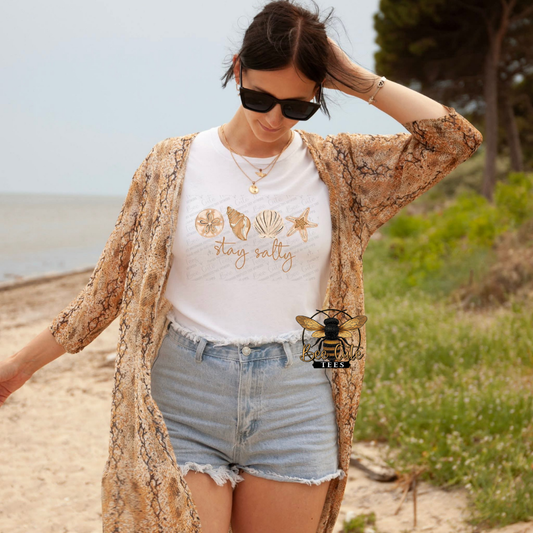 Woman on a beach wearing a white t-shirt with graphics and text, denim shorts, and a patterned cardigan.