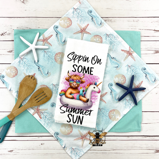 Tea towel with a cow on a unicorn and text 'Sippin On Some Summer Sun' on a decorative cloth with starfish and beach-themed items.