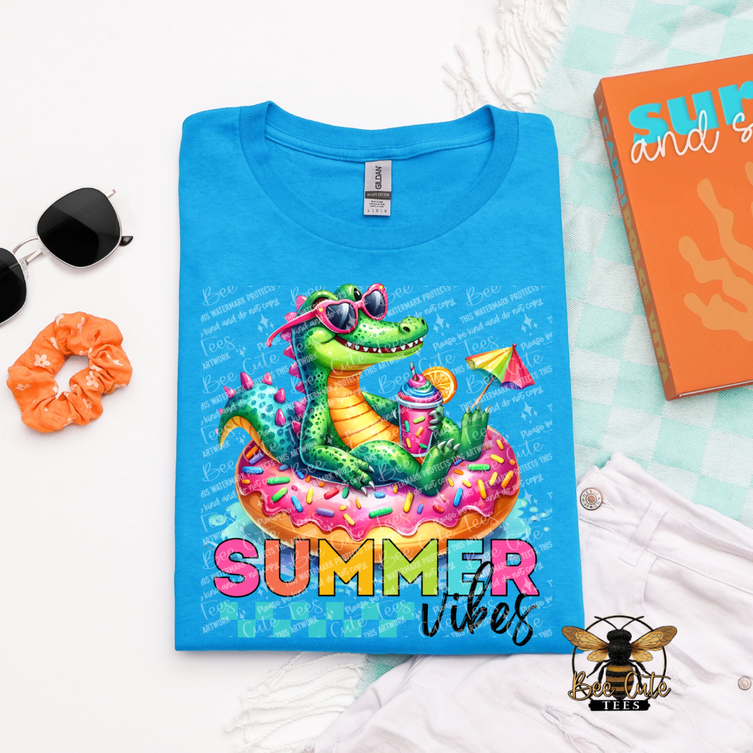 Blue t-shirt with a colorful alligator design and 'Summer Vibes' text, surrounded by sunglasses, scrunchie, and notebook.