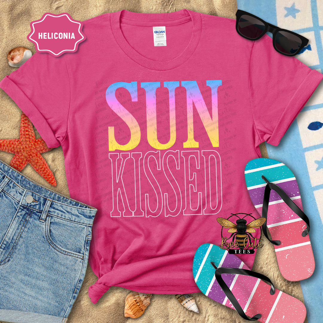 Pink t-shirt with 'SUN KISSED' text on a beach-themed background with sunglasses, flip-flops, and denim shorts.