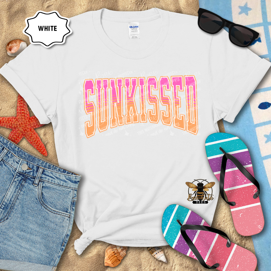 White t-shirt with 'SUNKISSED' text, sunglasses, denim shorts, and flip-flops on a sandy background.