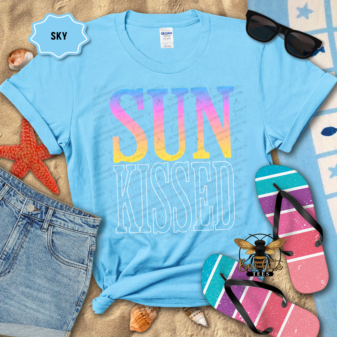 Blue t-shirt with 'SUN KISSED' text, sunglasses, flip-flops, and beach accessories on a sandy background.