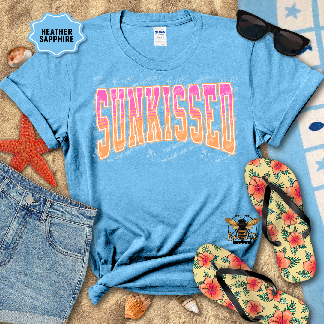 Blue t-shirt with 'SUNKISSED' text, sunglasses, flip-flops, and denim shorts on a sandy background.
