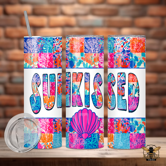 Bright and colorful 20 oz tumbler featuring a vibrant coral reef patchwork design with bold “Sunkissed” lettering and a pink seashell accent, displayed with a lid and metal straw against a rustic brick background.
