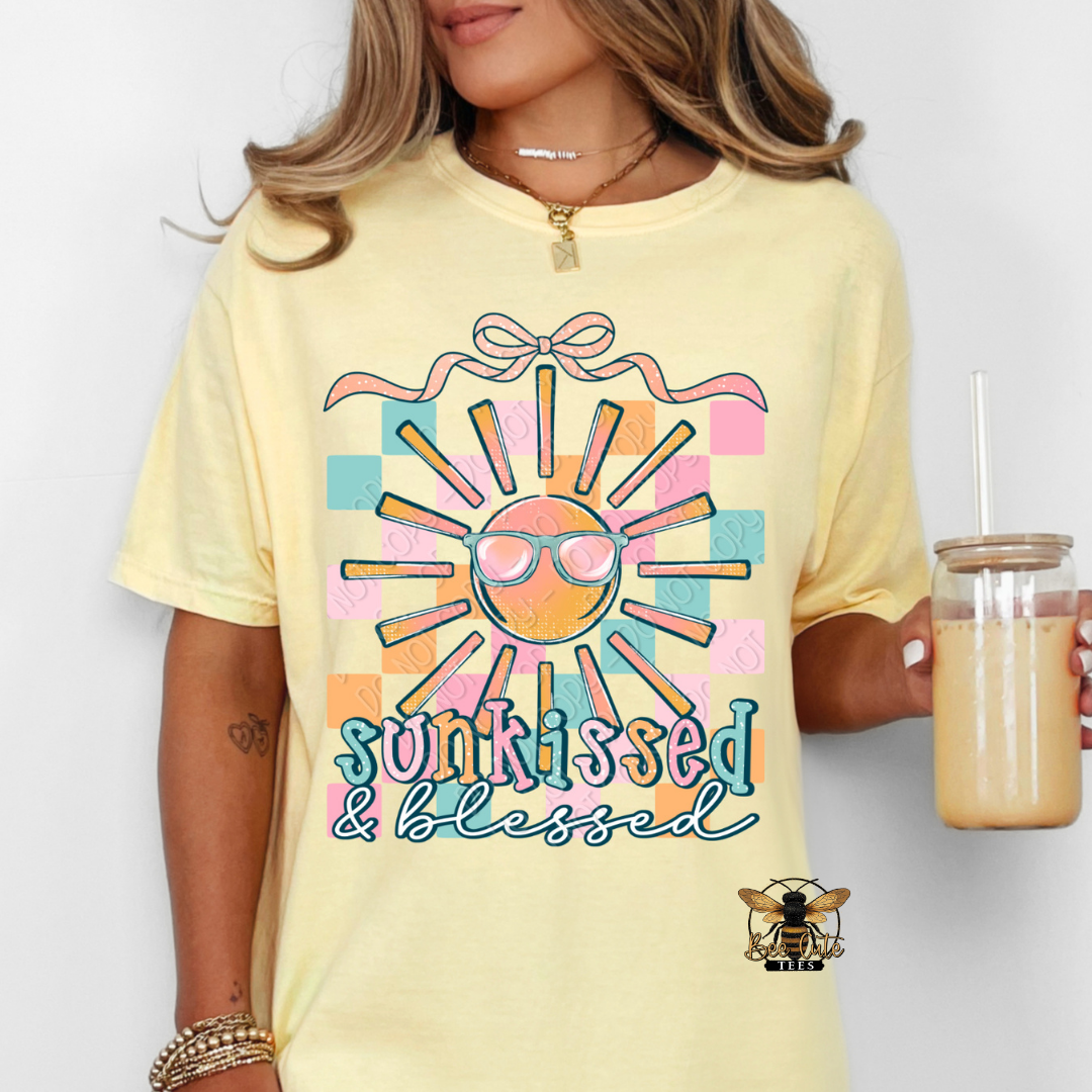 Person wearing a yellow t-shirt with a sun graphic and 'sunkissed & blessed' text, holding a drink.