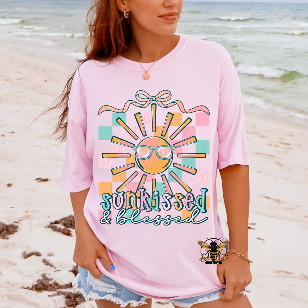 Person wearing a pink t-shirt with a sun graphic and 'sunkissed & blessed' text on a beach.