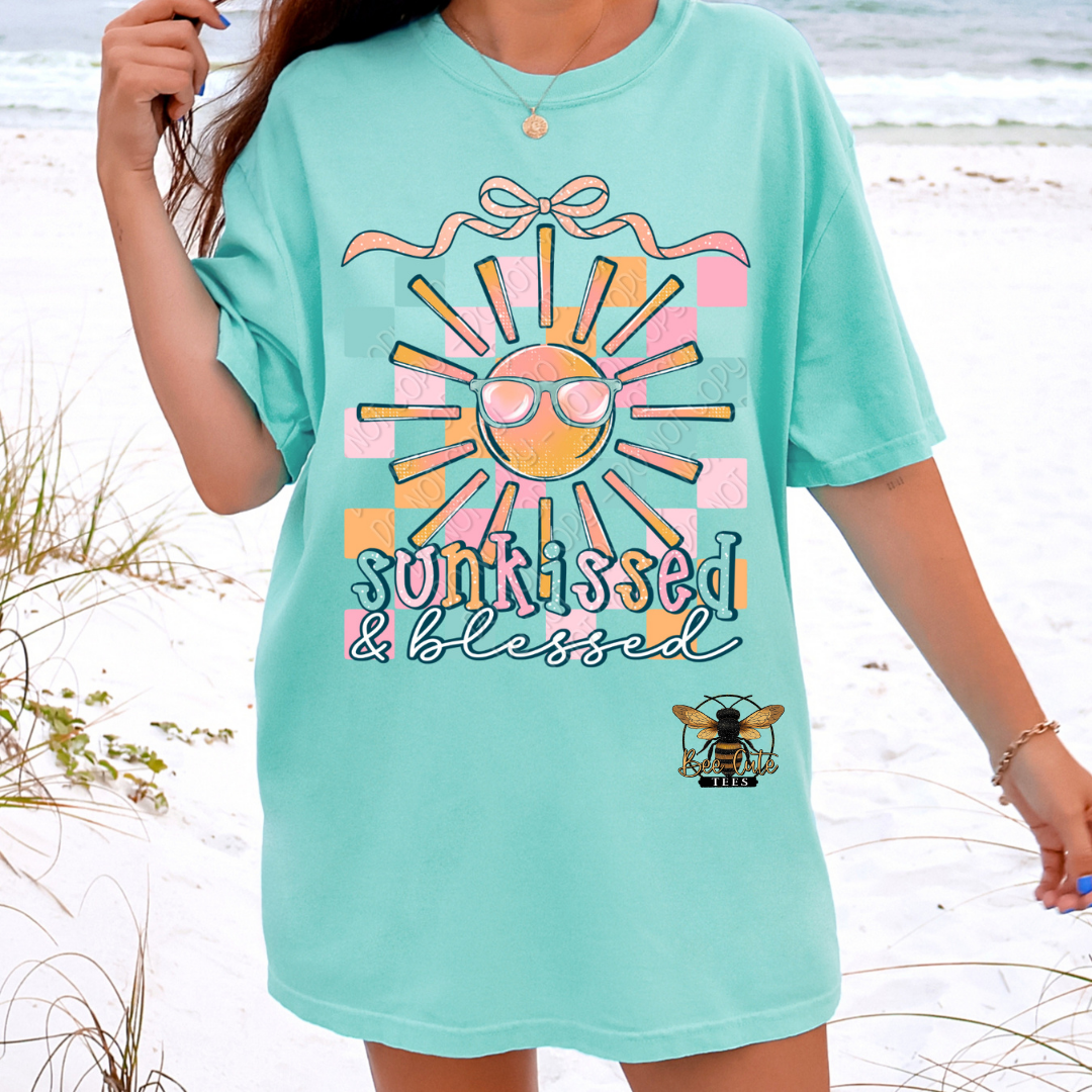 Person wearing a light blue t-shirt with a sun graphic and 'sunkissed & blessed' text on a beach.