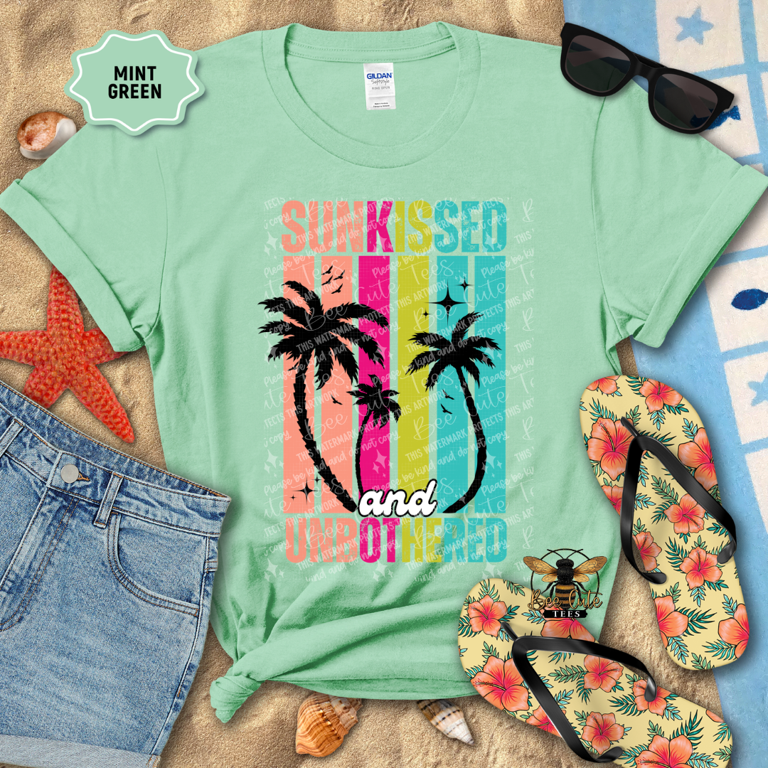 Mint green t-shirt with palm tree design and text, surrounded by beach accessories on a sandy background.