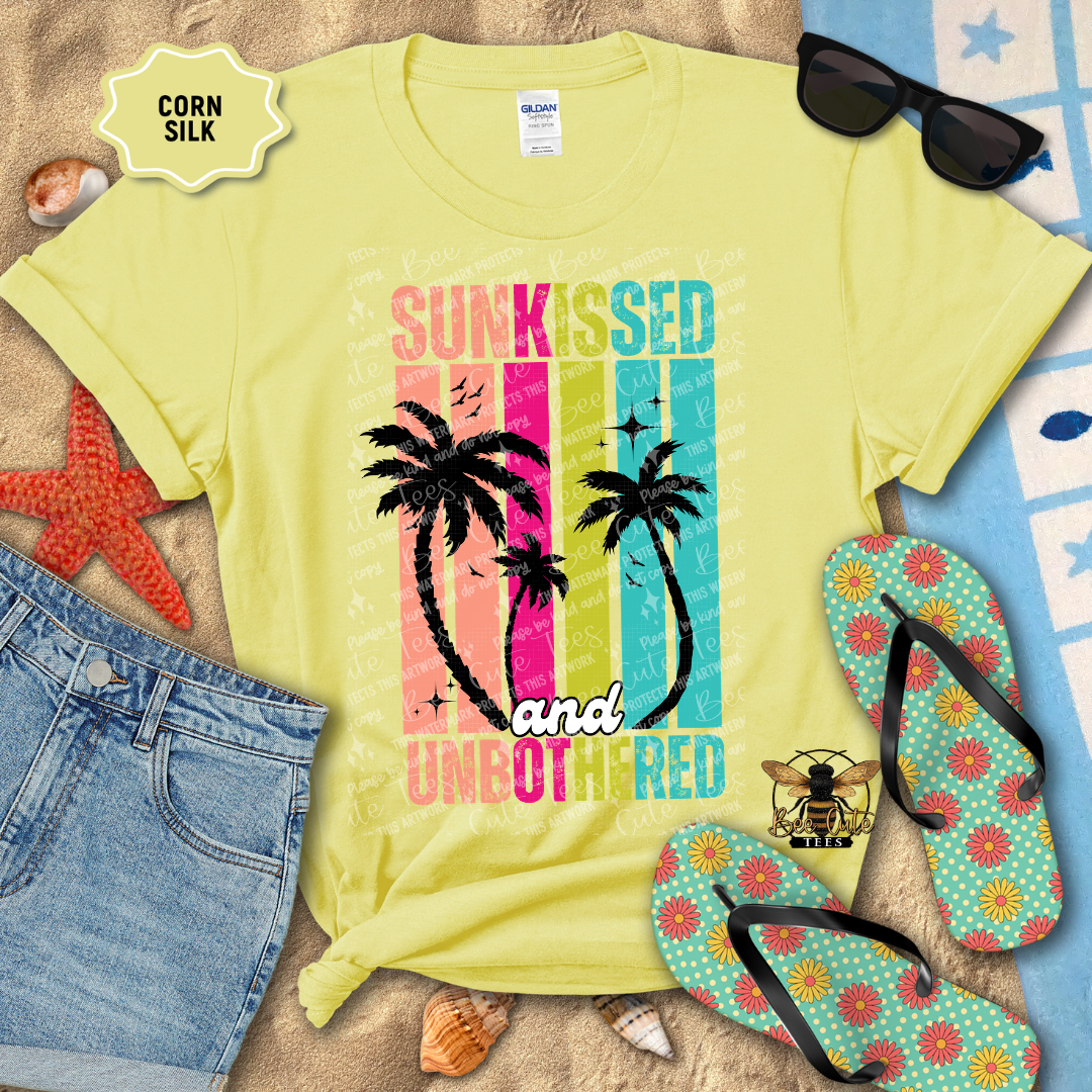 Yellow t-shirt with 'Sunkissed and Unbothered' design, sunglasses, flip-flops, and beach accessories on a sandy background.