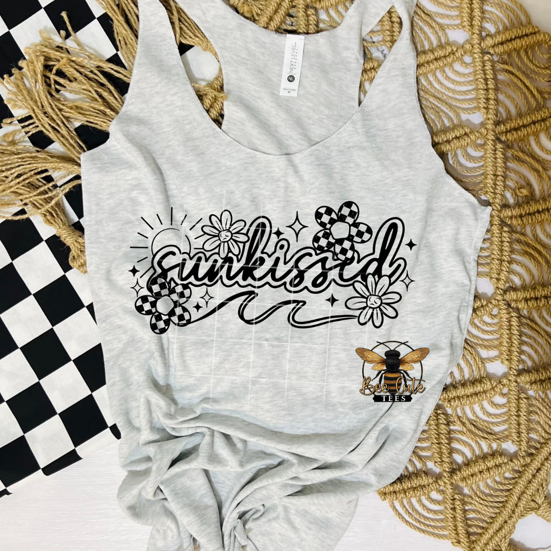 Light gray tank top with a black “Sunkissed” graphic in a playful script, surrounded by doodle-style flowers, sparkles, and a sun design. The shirt is styled flat lay on a woven mat with a black-and-white checkered accessory, and the Bee Cute Tees logo is visible.
