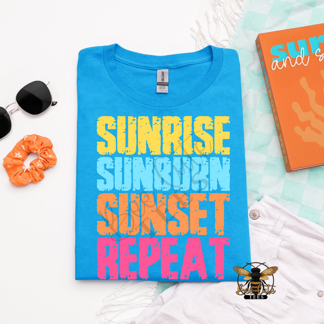 Blue t-shirt with 'Sunrise Sunburn Sunset Repeat' text on a white surface with accessories.