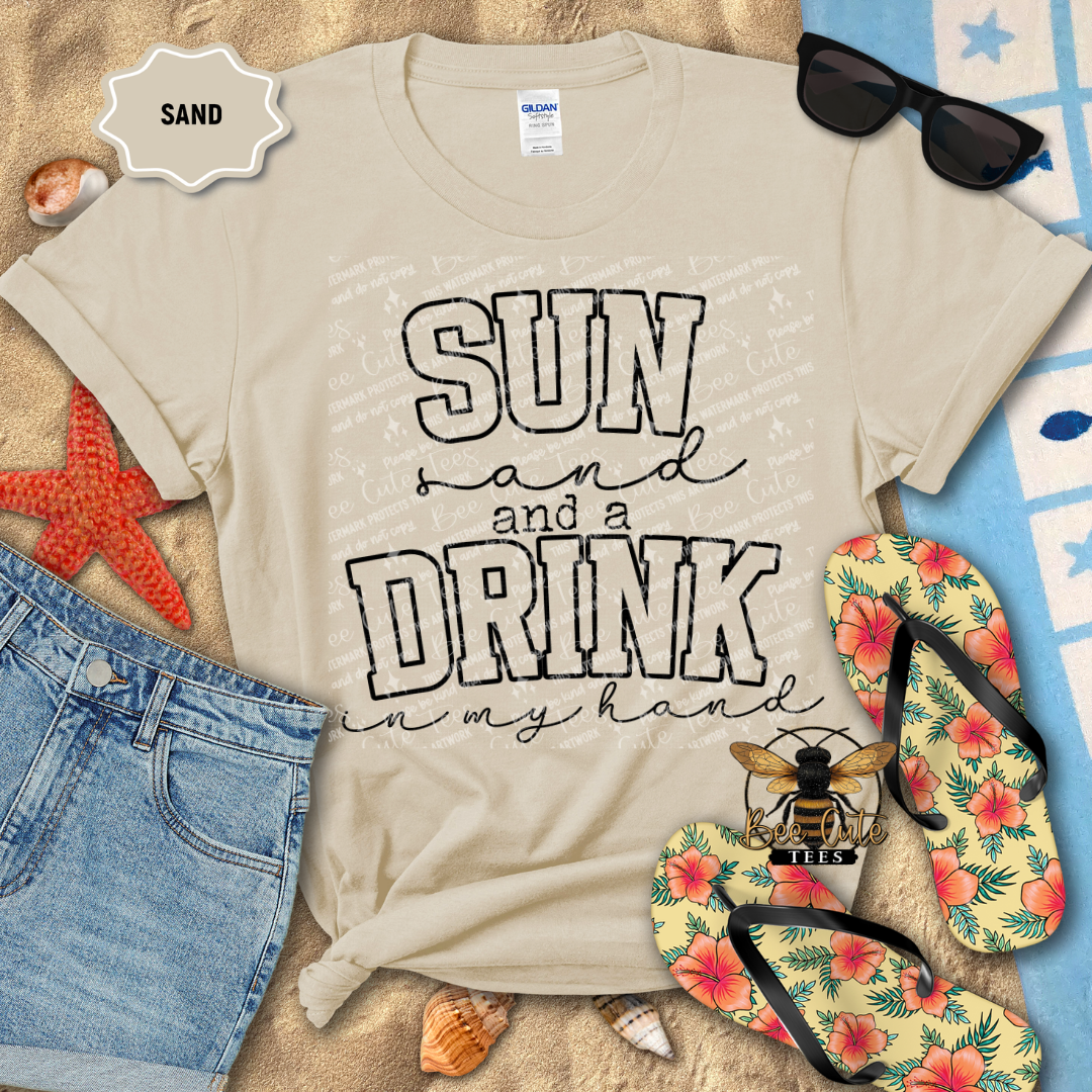 Beige t-shirt with 'Sun and a Drink in my hand' text, sunglasses, flip-flops, and beach items on sand.