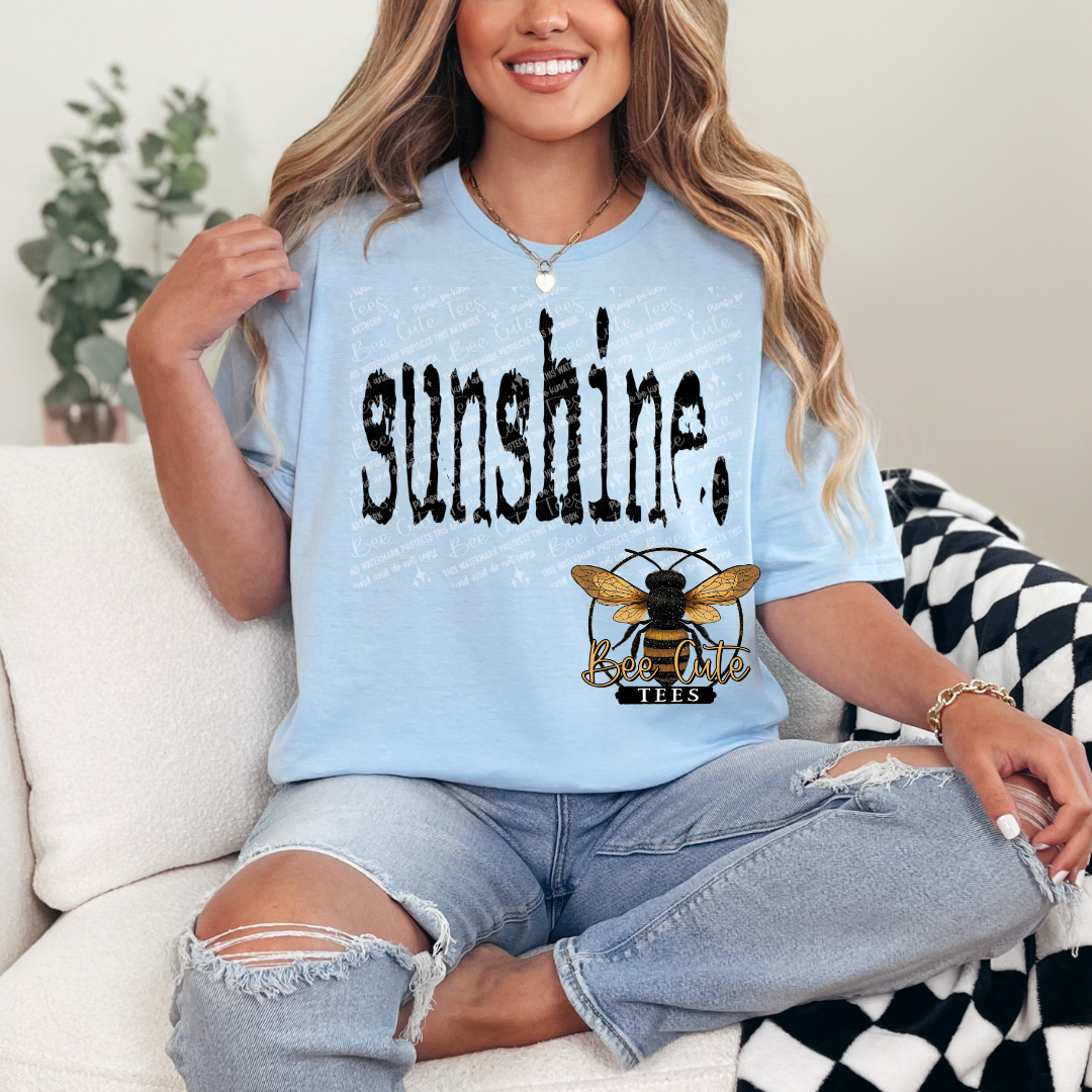 Person wearing a light blue t-shirt with 'sunshine' and a bee logo, sitting on a couch.