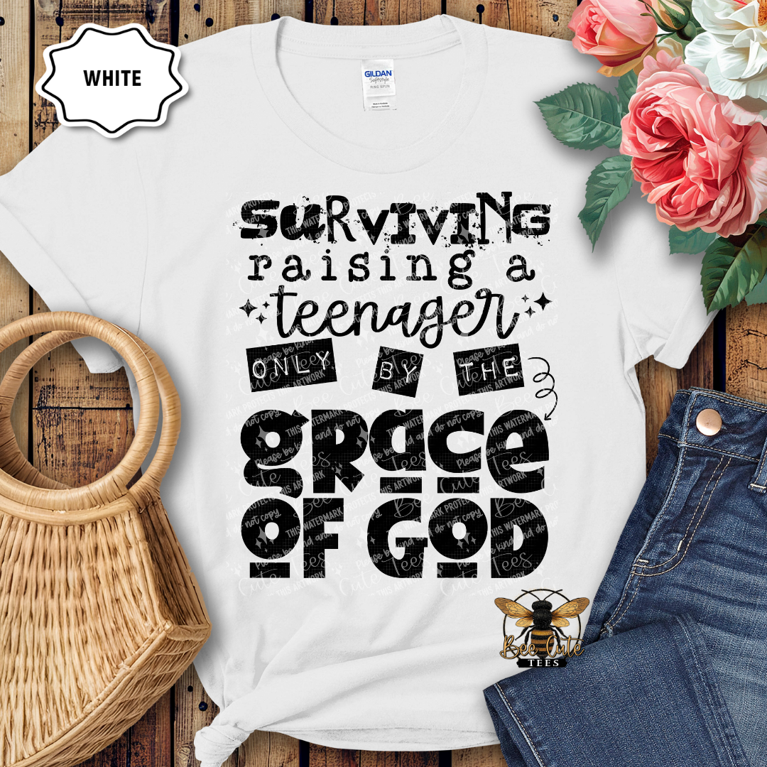 White t-shirt with black text 'Surviving raising a teenager only by the grace of God' on a wooden surface with flowers and jeans.