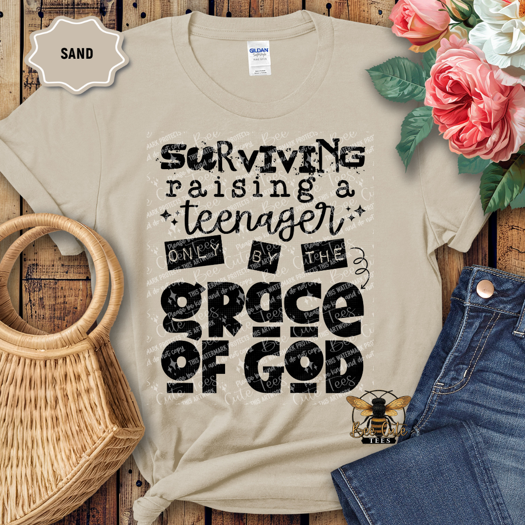 Beige t-shirt with text 'Surviving raising a teenager only by the grace of God' on a wooden surface with flowers and jeans.