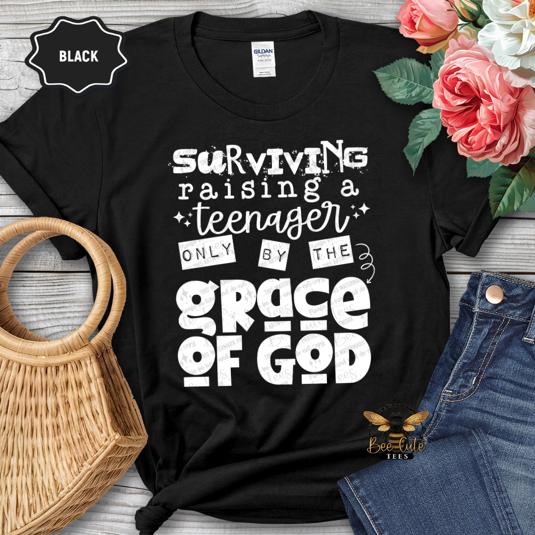 Black t-shirt with white text 'Surviving raising a teenager only by the grace of God' on a wooden surface with flowers and jeans.