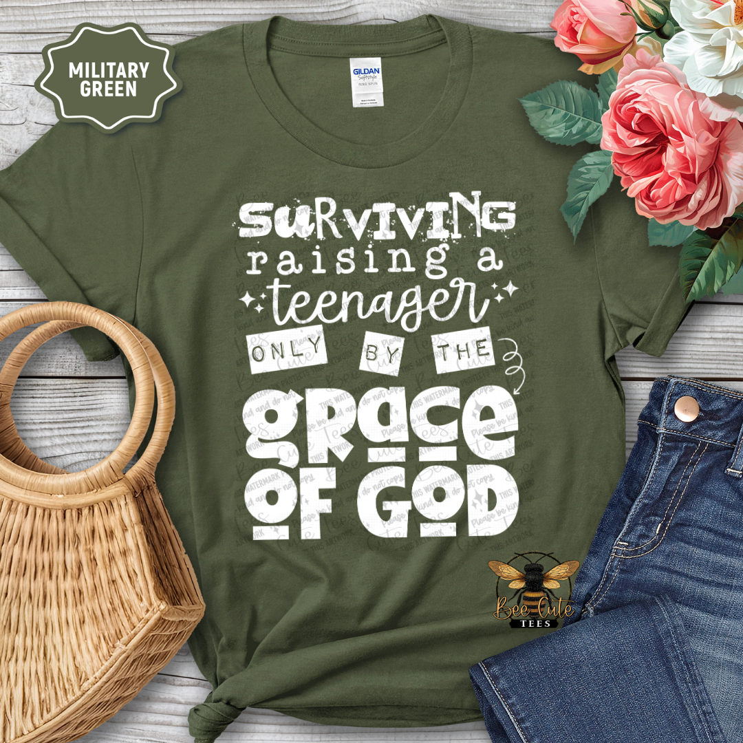 Green t-shirt with text 'Surviving raising a teenager only by the grace of God' on a wooden surface with flowers and jeans.