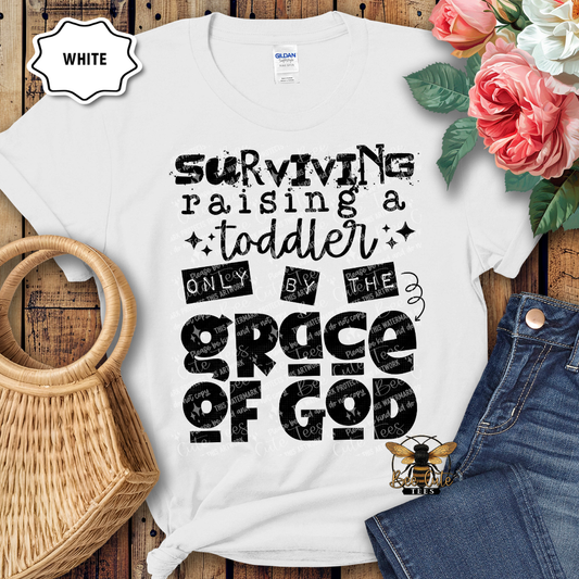 White t-shirt with black text 'Surviving raising a toddler only by the grace of God' on a wooden surface with flowers and jeans.