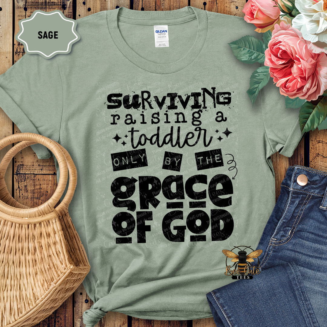 Sage green t-shirt with black text 'Surviving raising a toddler only by the grace of God' on a wooden surface with flowers and jeans.