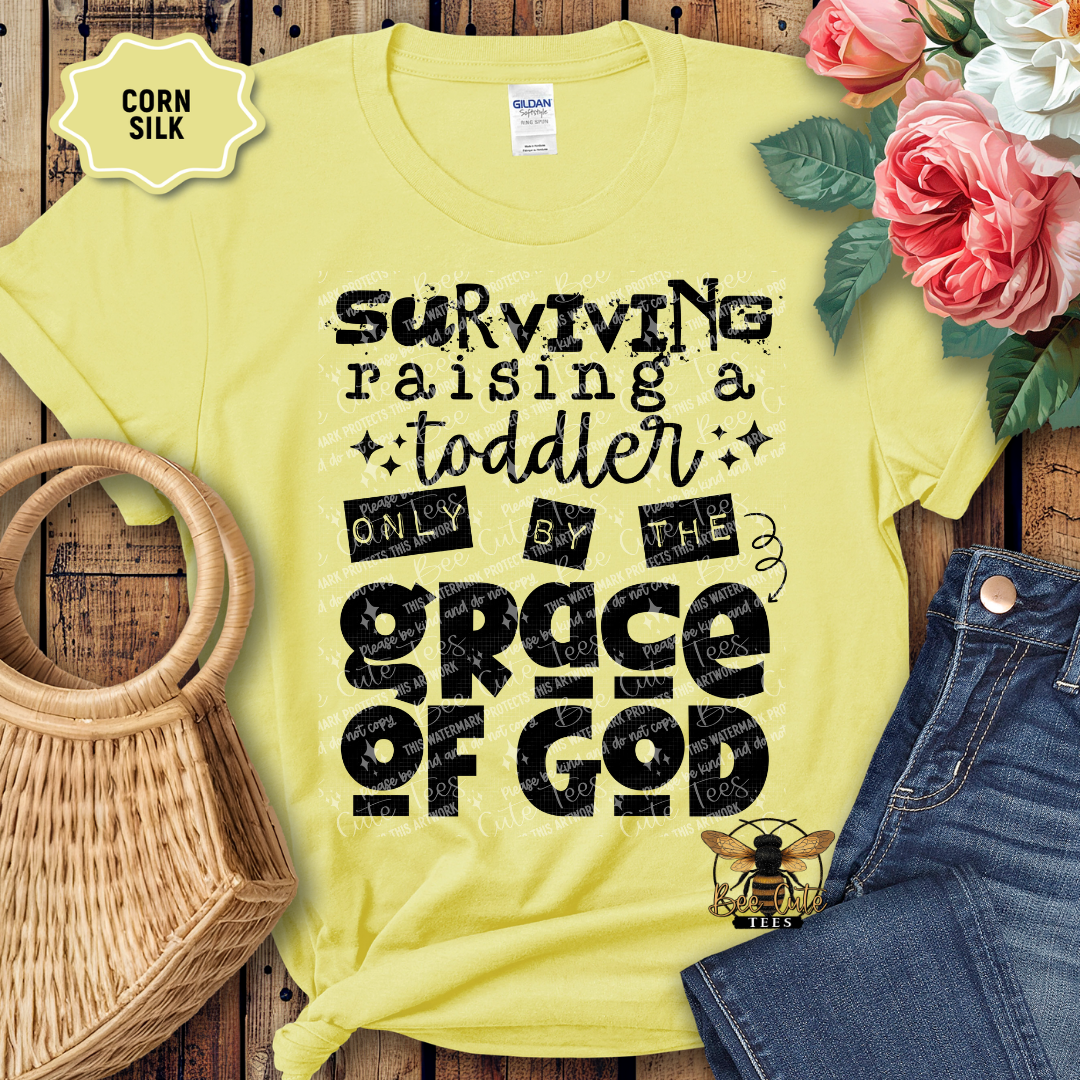 Yellow t-shirt with text 'Surviving raising a toddler only by the grace of God' on a wooden surface with flowers and jeans.