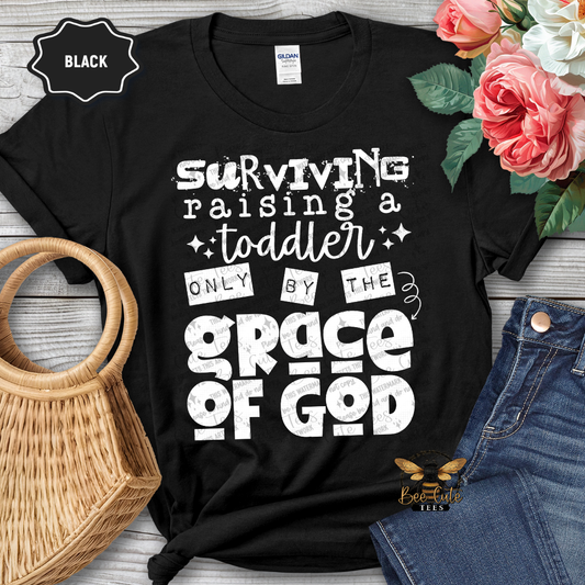 Black t-shirt with white text 'Surviving raising a toddler only by the grace of God' on a wooden surface with flowers and jeans.