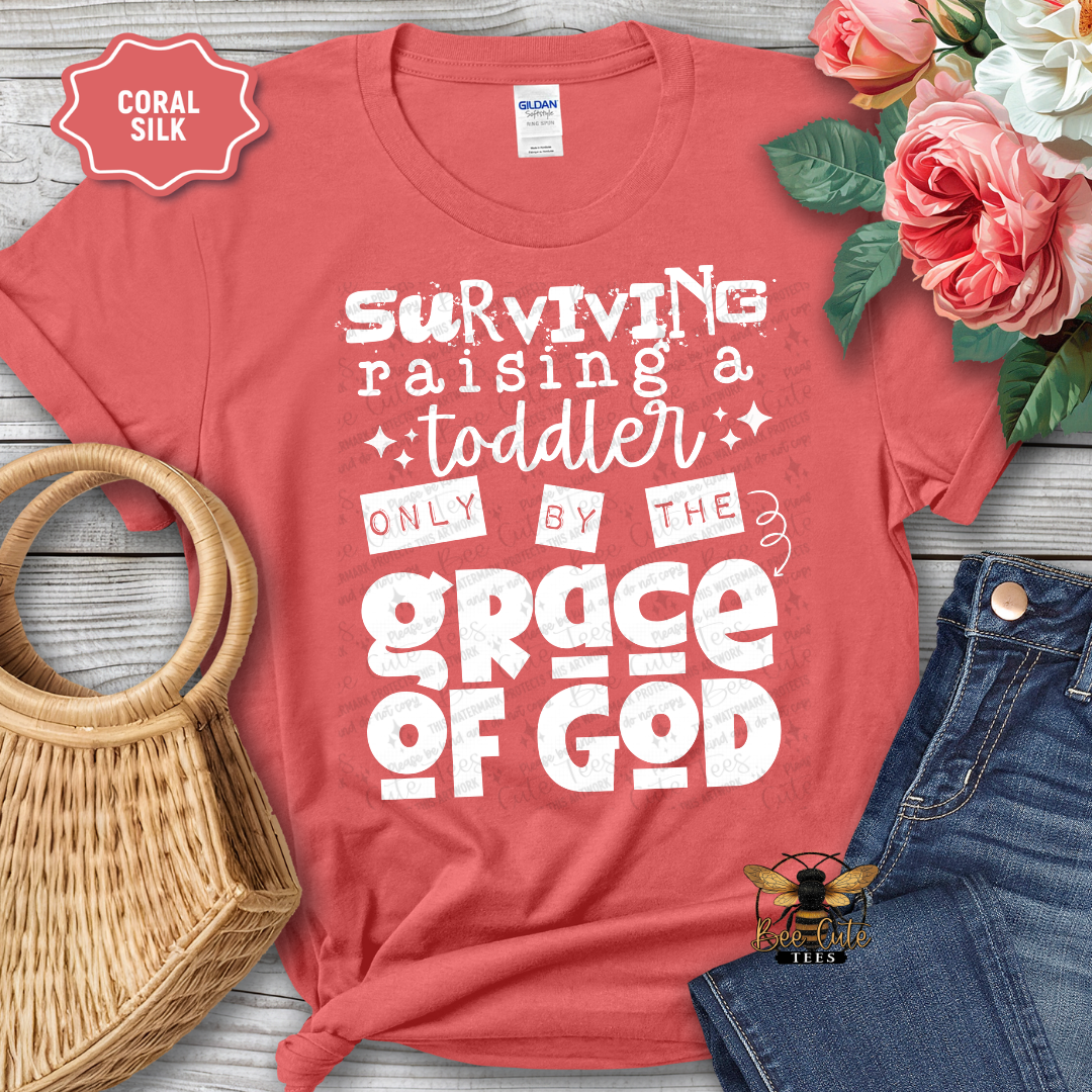 Coral t-shirt with text 'Surviving raising a toddler only by the grace of God' on a wooden surface with flowers and jeans.