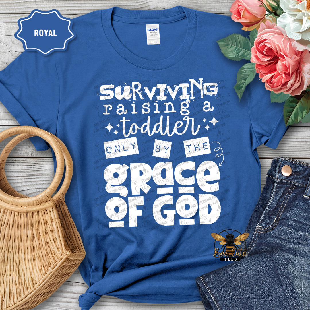 Blue t-shirt with white text 'Surviving raising a toddler only by the grace of God' on a wooden surface with flowers and a bag.