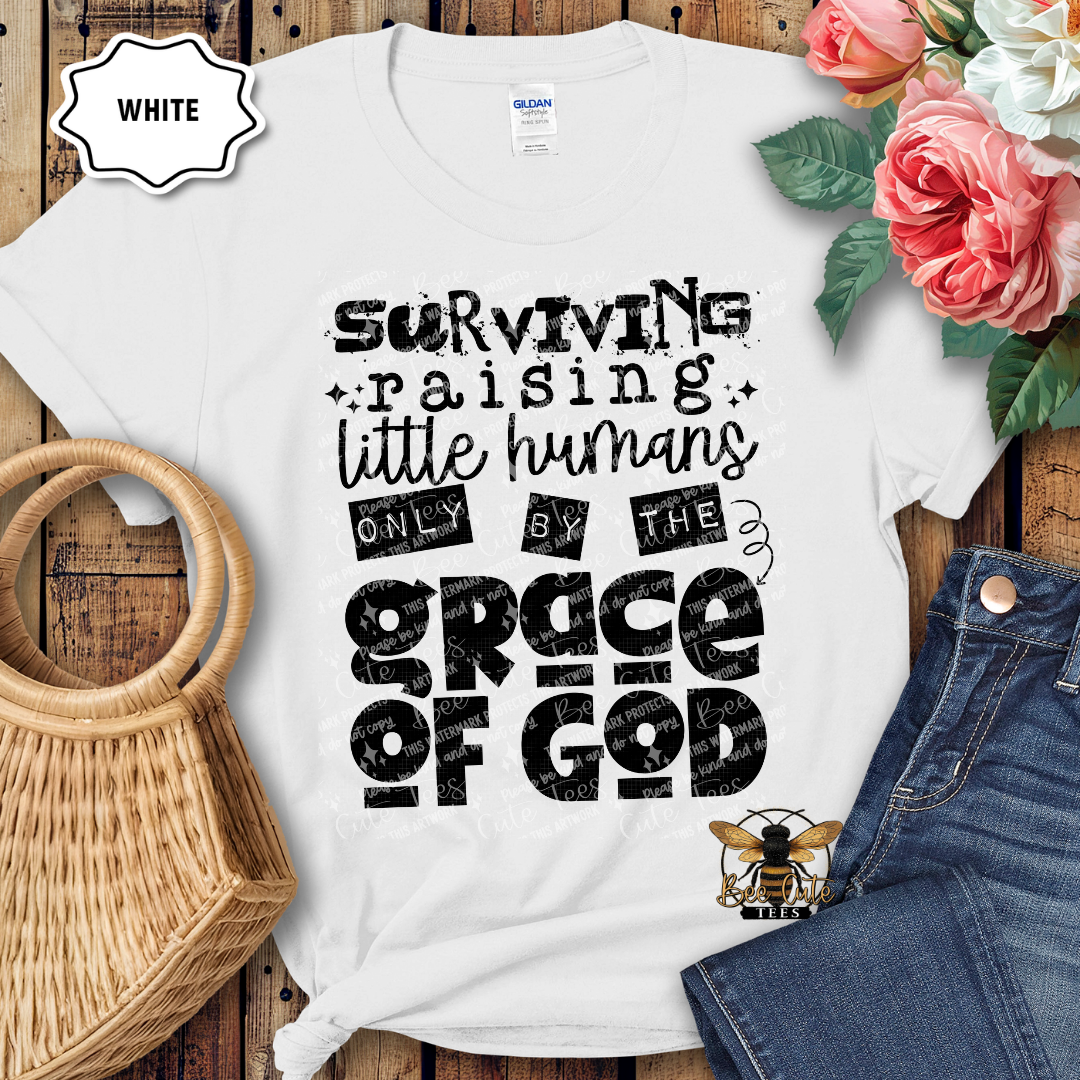 White t-shirt with black text 'Surviving raising little humans only by the grace of God' on a wooden surface with flowers and jeans.