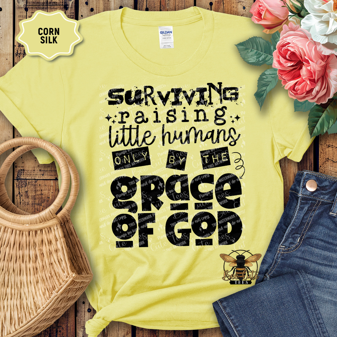 Yellow t-shirt with text 'Surviving raising little humans only by the grace of God' on a wooden surface with flowers and jeans.