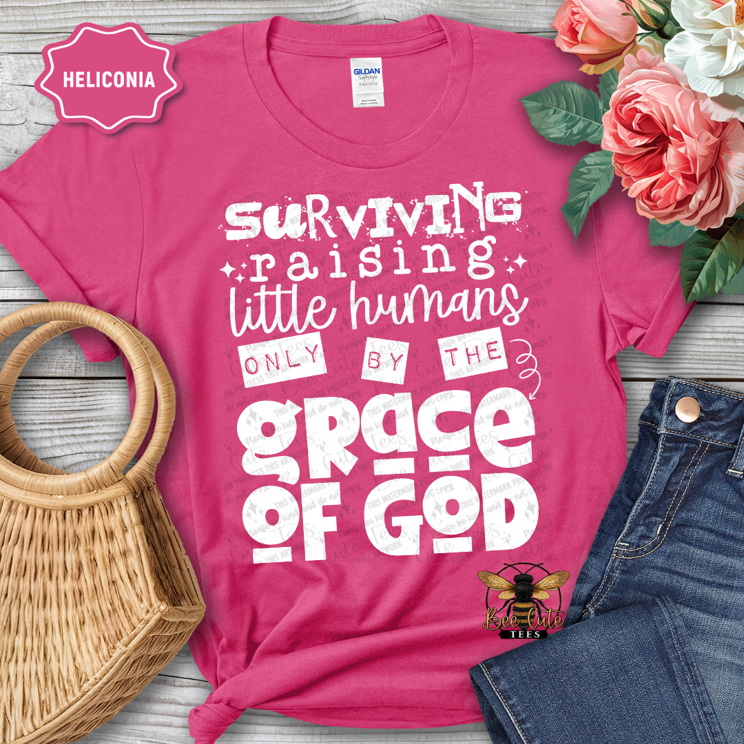 Pink t-shirt with white text 'Surviving raising little humans only by the grace of God' on a wooden surface with flowers and jeans.