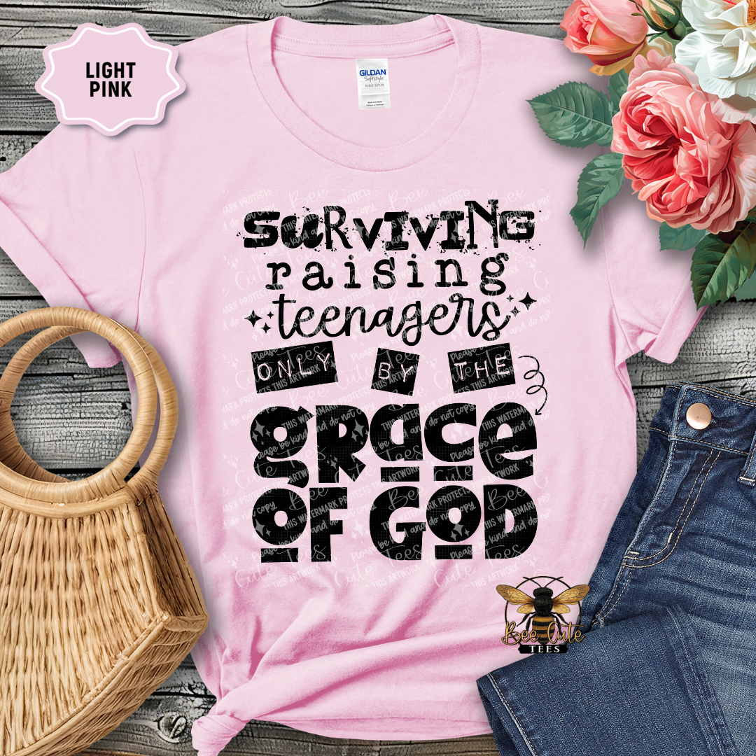 Pink t-shirt with text 'Surviving raising teenagers only by the grace of God' on a wooden surface with flowers and accessories.