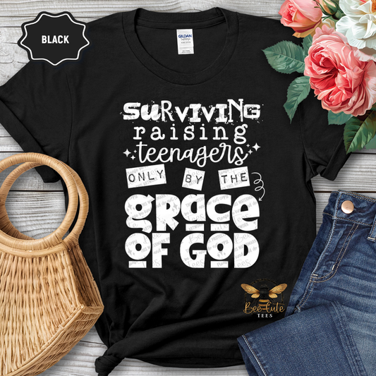Black t-shirt with white text 'Surviving raising teenagers only by the grace of God' on a wooden surface with flowers and jeans.