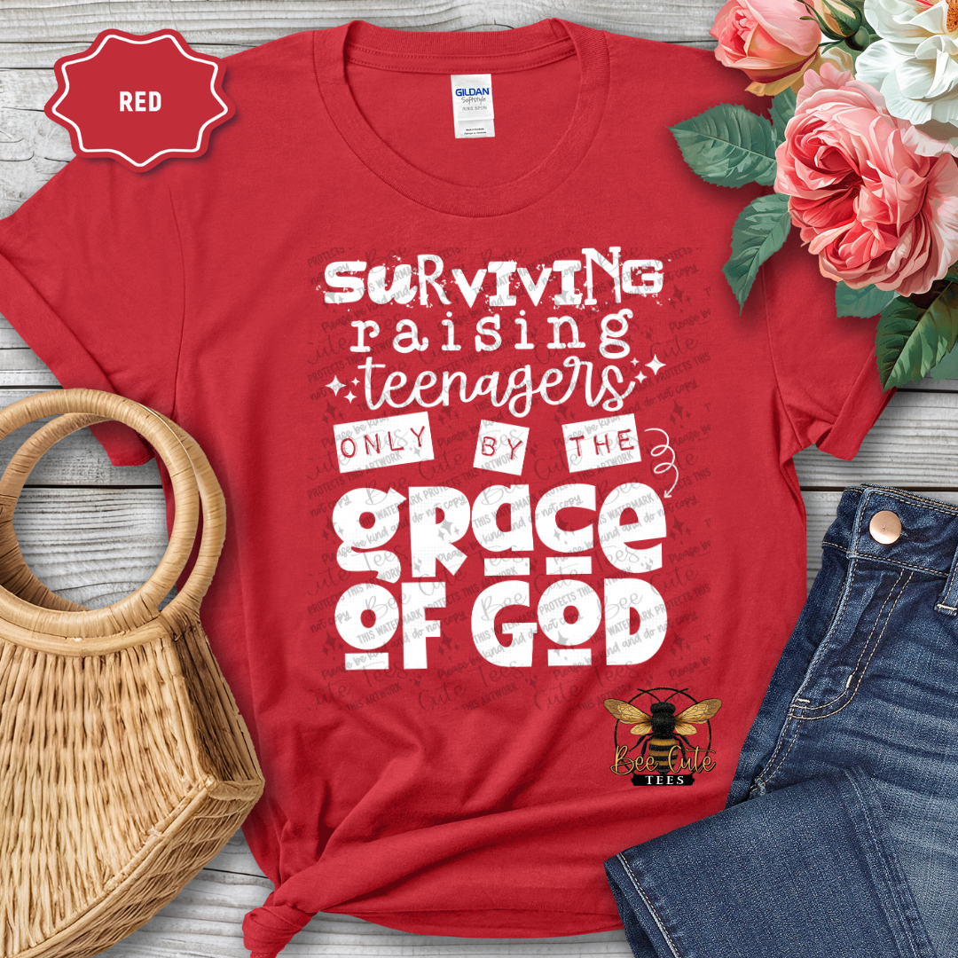 Red t-shirt with text 'Surviving raising teenagers only by the grace of God' on a wooden surface with flowers and jeans.