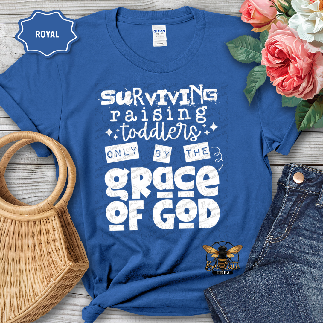 Blue t-shirt with white text 'Surviving raising toddlers only by the grace of God' on a wooden surface with flowers and jeans.
