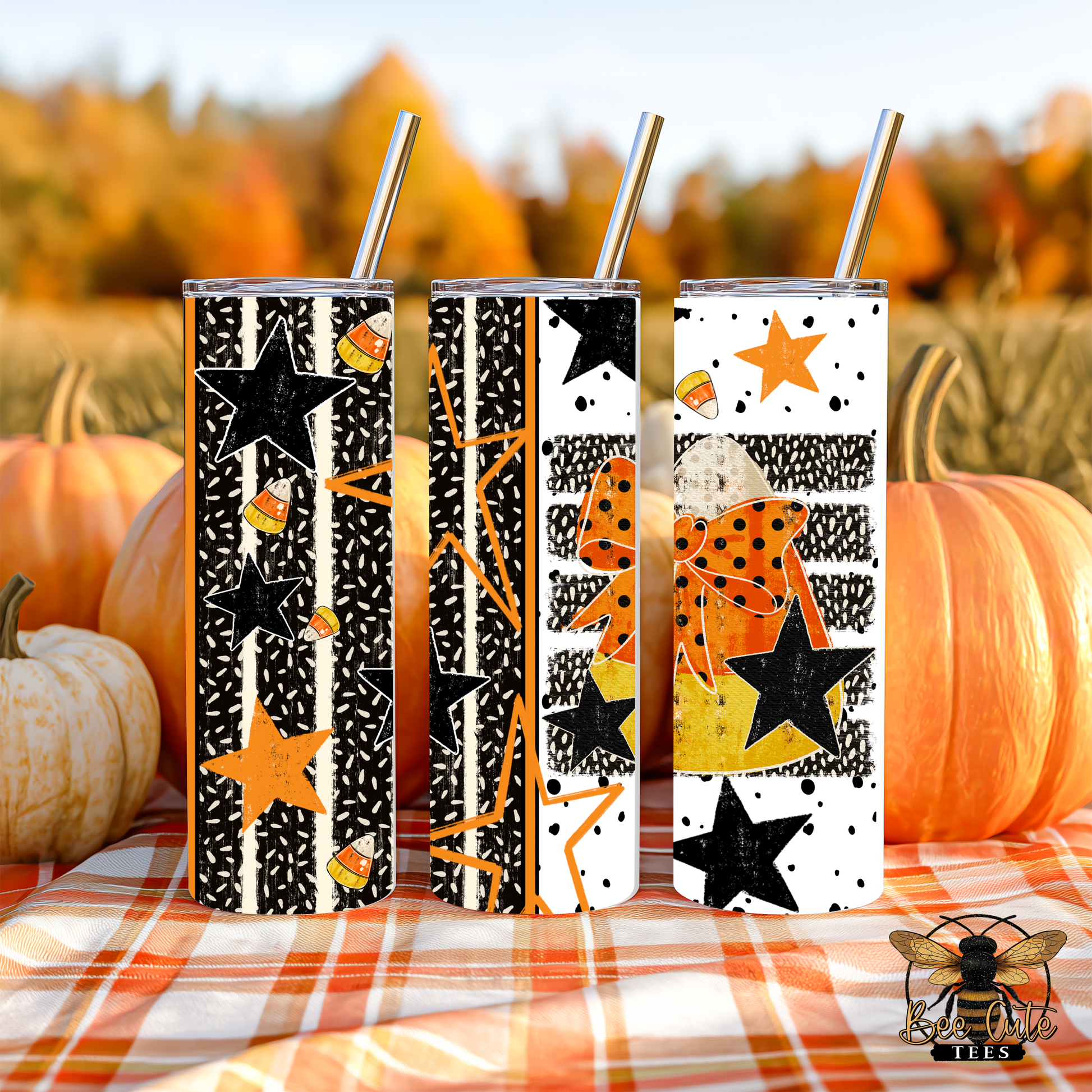 Three insulated tumblers with metal straws are displayed outdoors on an orange plaid blanket with pumpkins and a blurred autumn field behind them. The tumblers feature black, white, and orange fall-themed patterns including stars, polka dots, stripes, and candy corn motifs.