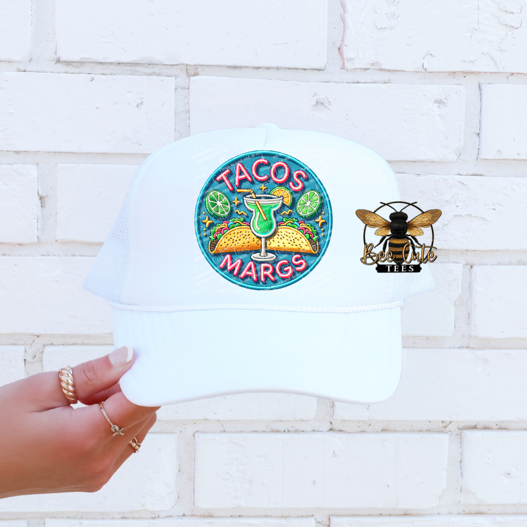 White t-shirt with 'TACOS MARGS' design held against a white brick wall.