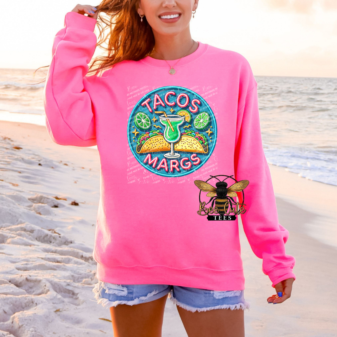 Person wearing a pink sweatshirt with 'Tacos Margs' design on a beach