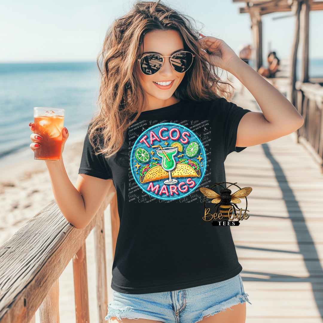 Woman wearing a black t-shirt with 'TACOS MARGS' design, standing on a wooden pier by the ocean.