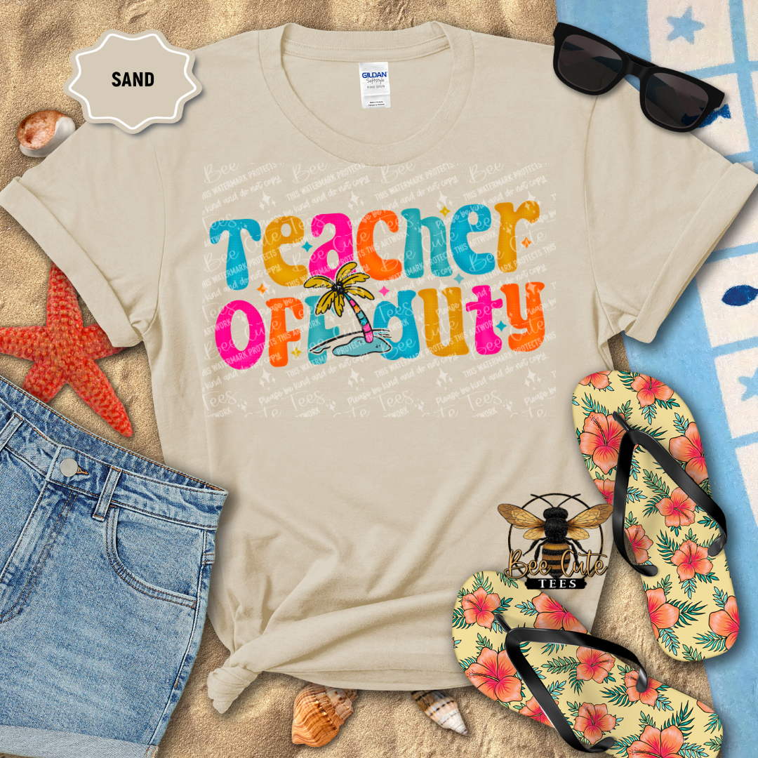 Beige t-shirt with 'Teacher Off Duty' text on a sandy background with beach accessories.