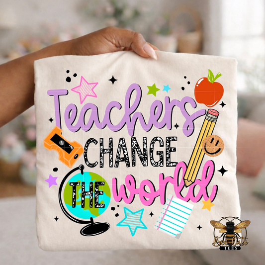 Folded white unisex T-shirt held up by a hand, featuring a colorful "Teachers Change the World" graphic. The design combines purple, black, and pink lettering with playful school-themed illustrations including a pencil, apple, globe, notebook paper, pencil sharpener, smiley face, and colorful stars. A fun, vibrant design perfect for teachers and educators.

