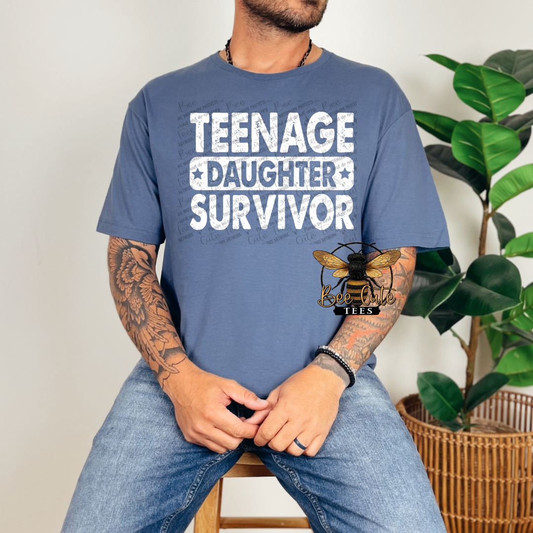 Person wearing a blue t-shirt with 'TEENAGE DAUGHTER SURVIVOR' text and a bee logo, sitting indoors.