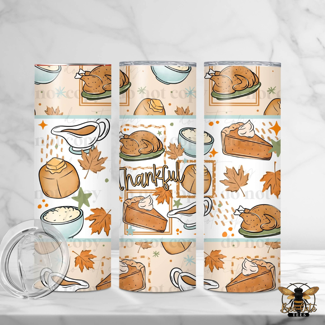 Three insulated tumblers with a Thanksgiving-themed pattern featuring roasted turkey, pie, pumpkin, maple leaves, gravy, and the word 'Thankful' on a light cream background. A clear lid is shown in the lower left on a white marble surface.